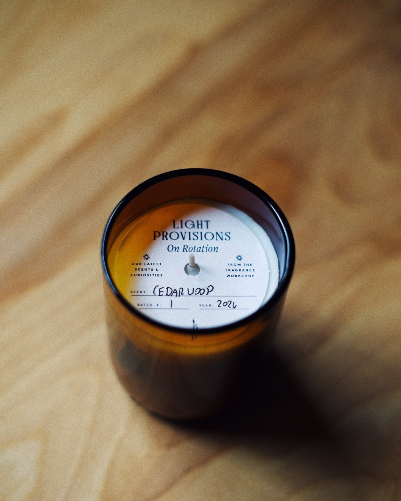 An amber glass jar candle with a white circular label reading 'LIGHT PROVISIONS On Rotation' and handwritten text, placed on a warm wood surface.