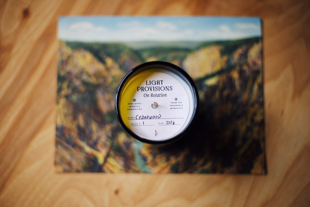 A circular candle jar with a yellow and white label reading 'LIGHT PROVISIONS On Rotation' sits on top of a square art print depicting a canyon landscape, all placed on a wooden surface.