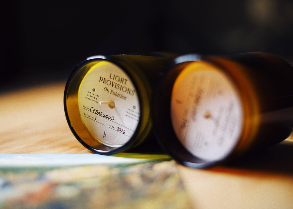 Two amber glass jar candles lying on their sides on a wooden surface, with one candle label clearly visible reading 'LIGHT PROVISIONS' and 'Cedarwood'.