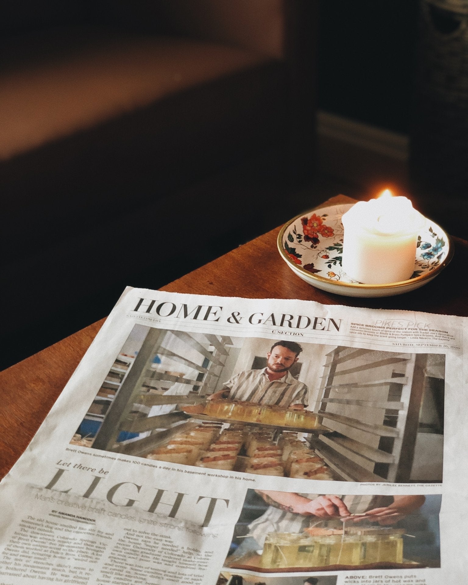 A newspaper page titled HOME & GARDEN lies on a wooden table next to a lit white candle in a small floral dish, with a dark indoor background.
