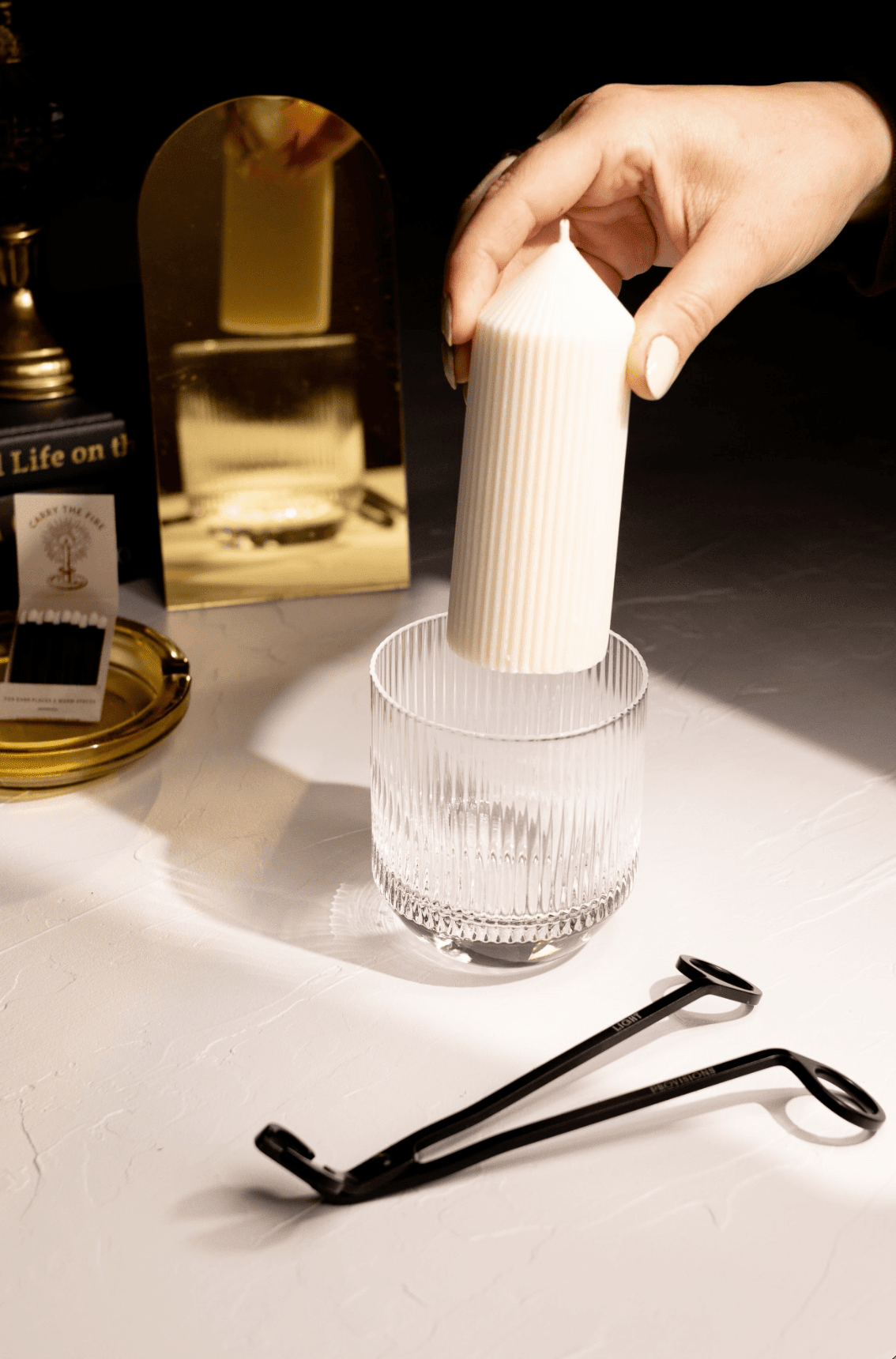 Hand placing a white refill candle into a glass tumbler on a reflective surface with a blurred background.