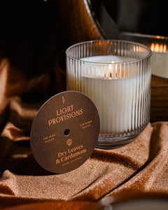 Light Provisions Dry Leaves & Cardamom candle in signature glass tumbler with branded dust cover on warm backdrop