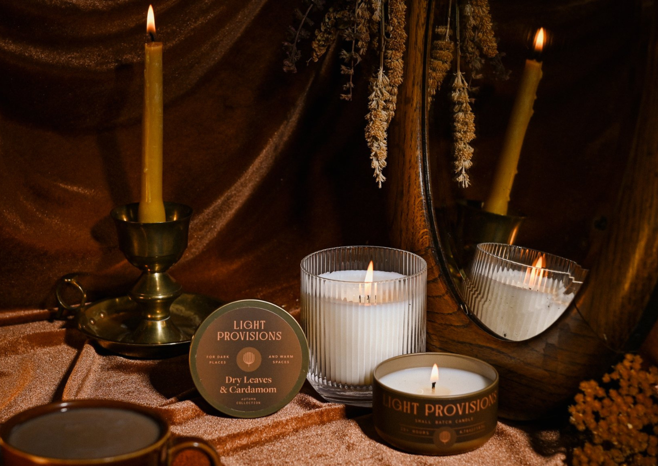 A styled product photo of Light Provisions candles. The set includes a tin candle labeled 'Dry Leaves & Cardamom', a jar candle in a ribbed glass holder, and a smaller tin candle, all lit and arranged on a draped brown fabric surface next to brass candlesticks and dried flowers.