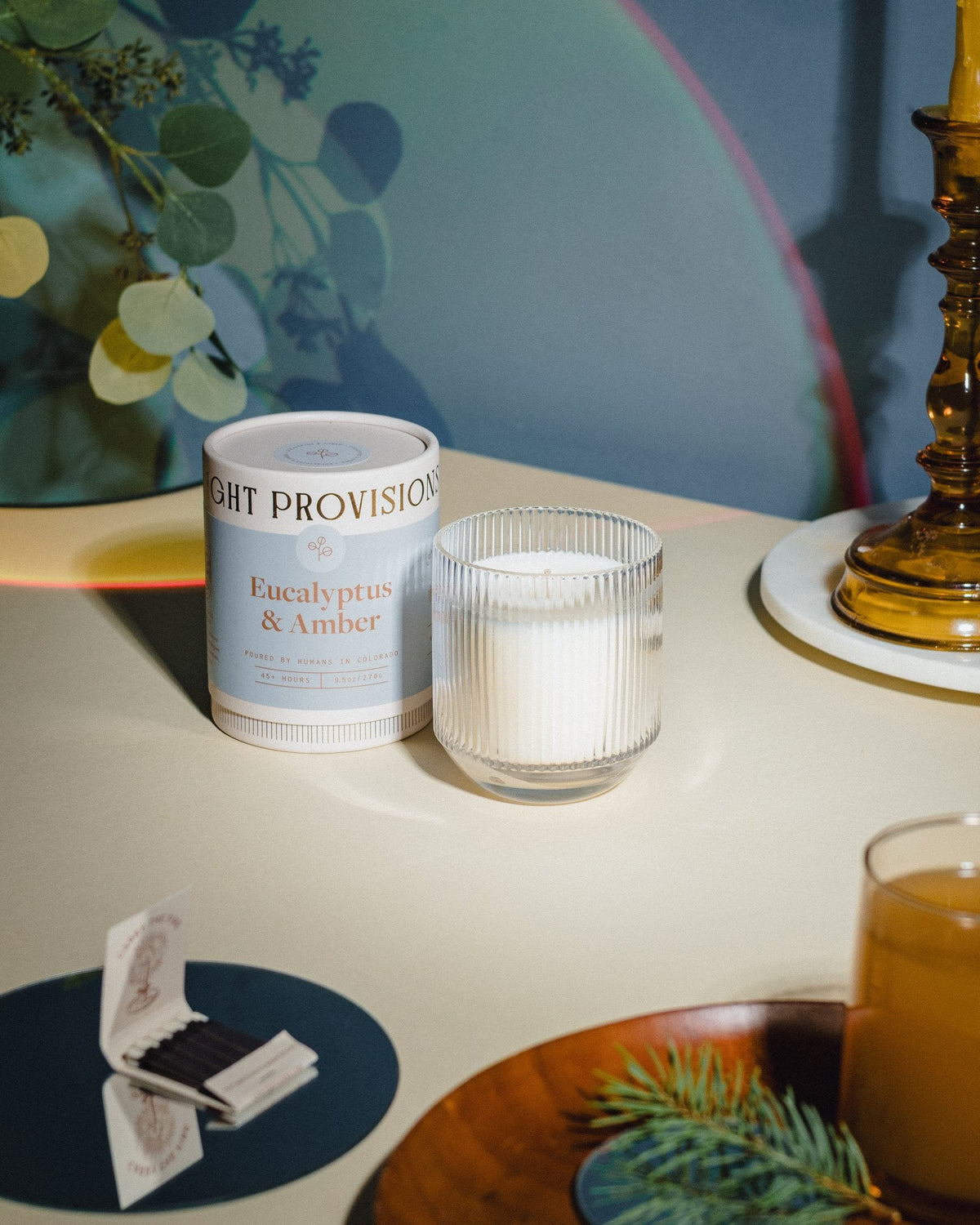 Eucalyptus & Amber candles with brass candlestick, drink glass, and greenery reflected in soft blue rainbow on wall