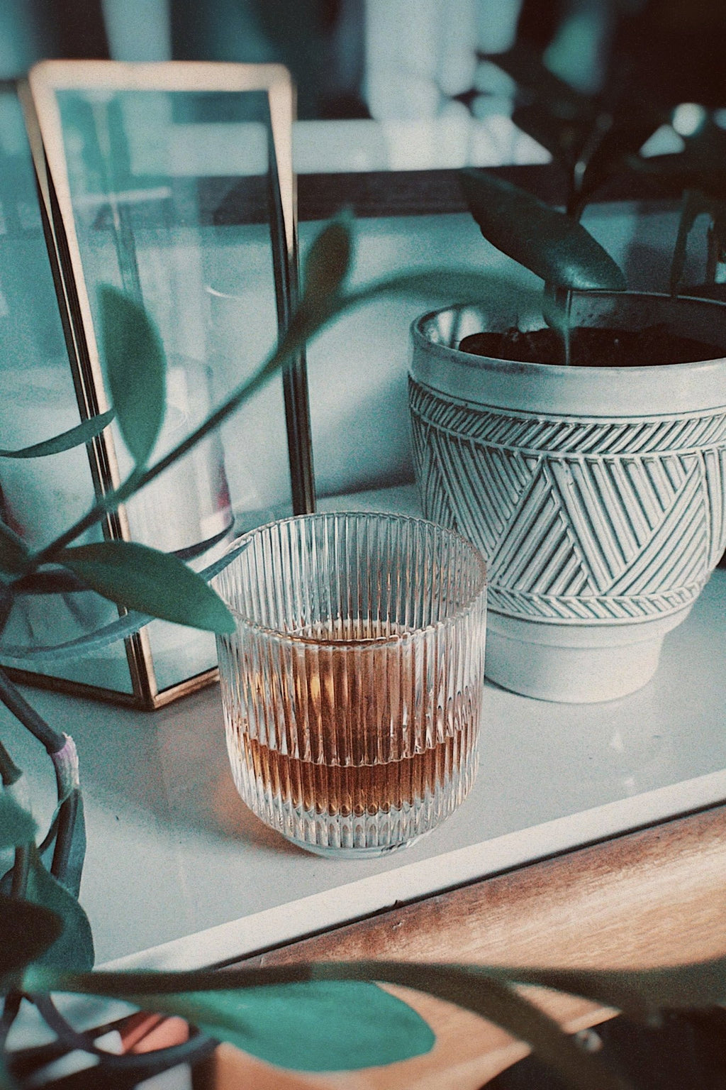 Ripple glassware with an amber cocktail, photographed on a shelf beside houseplants, a lantern, and a ceramic planter.