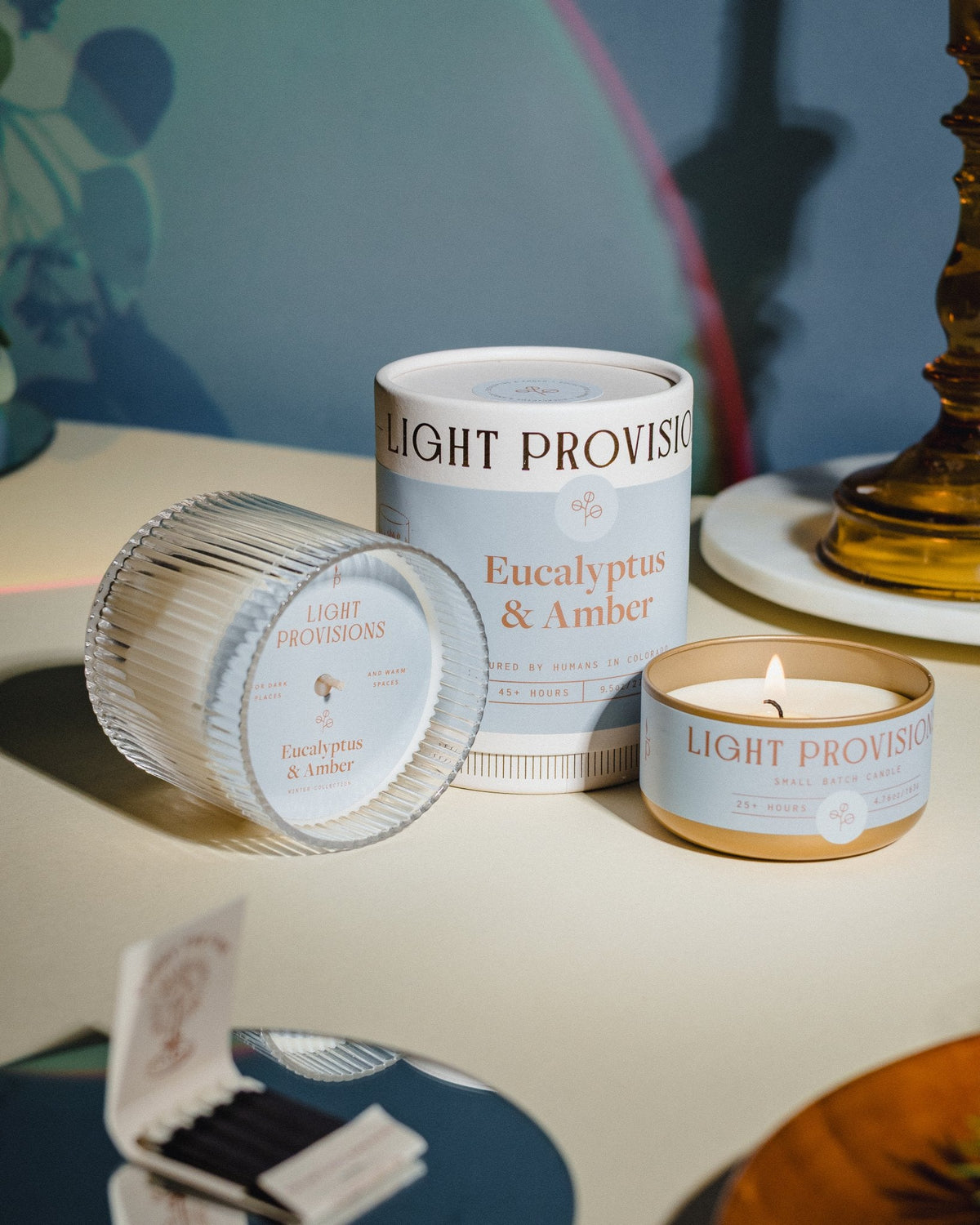Eucalyptus & Amber candle with packaging and ribbed tin beside a brass candlestick and green foliage casting soft shadows.