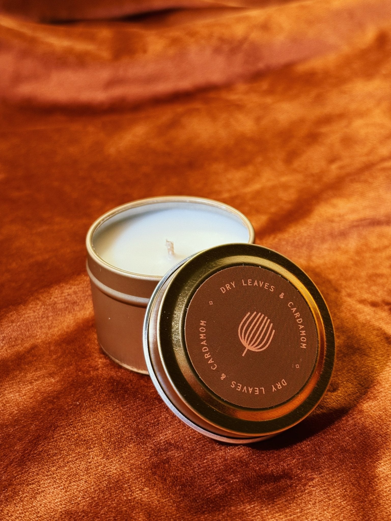 Dry Leaves Cardamom 2 oz tin sample candle with brown lid on burnt orange fabric background