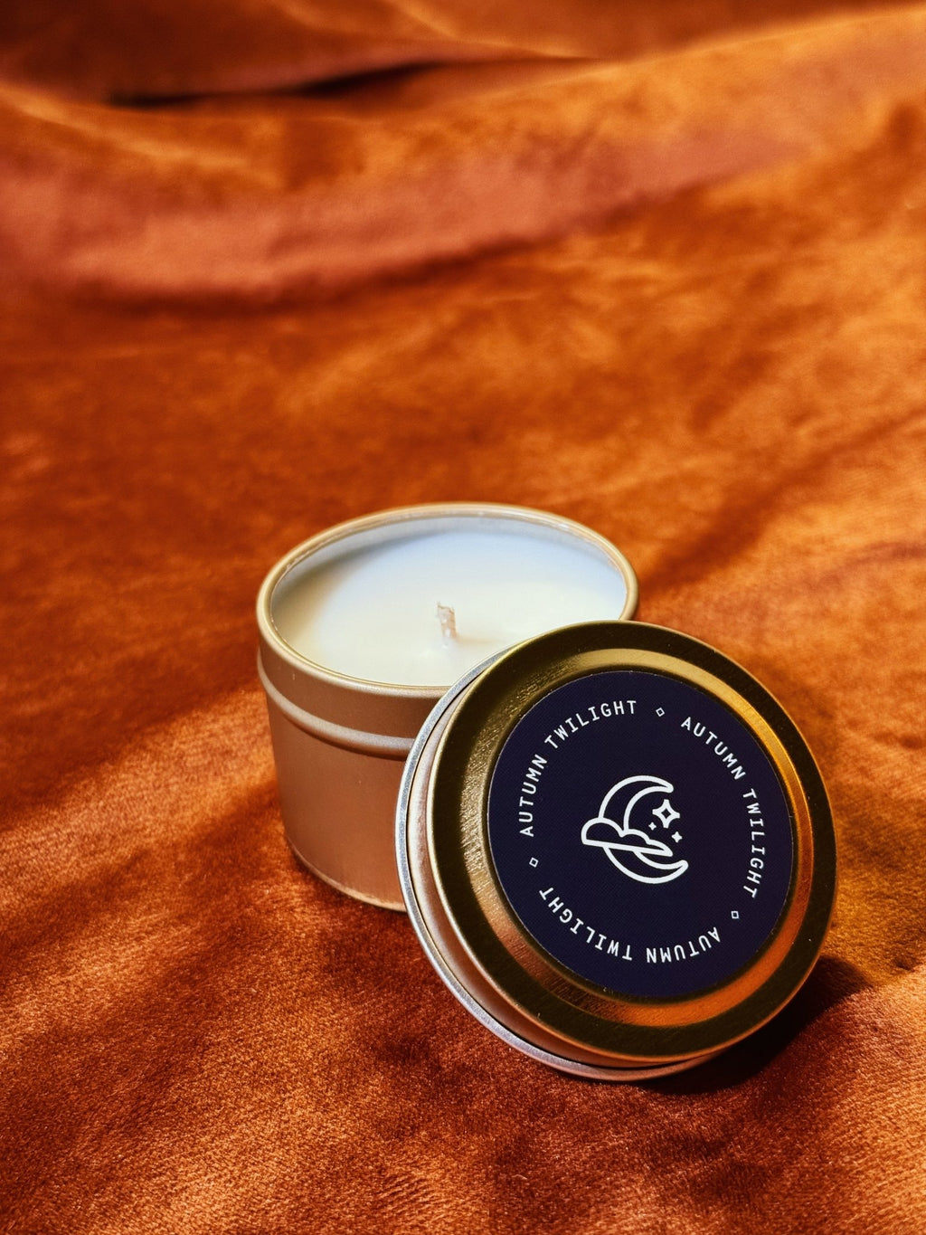 Autumn Twilight 2 oz tin sample candle with dark blue lid on burnt orange fabric background