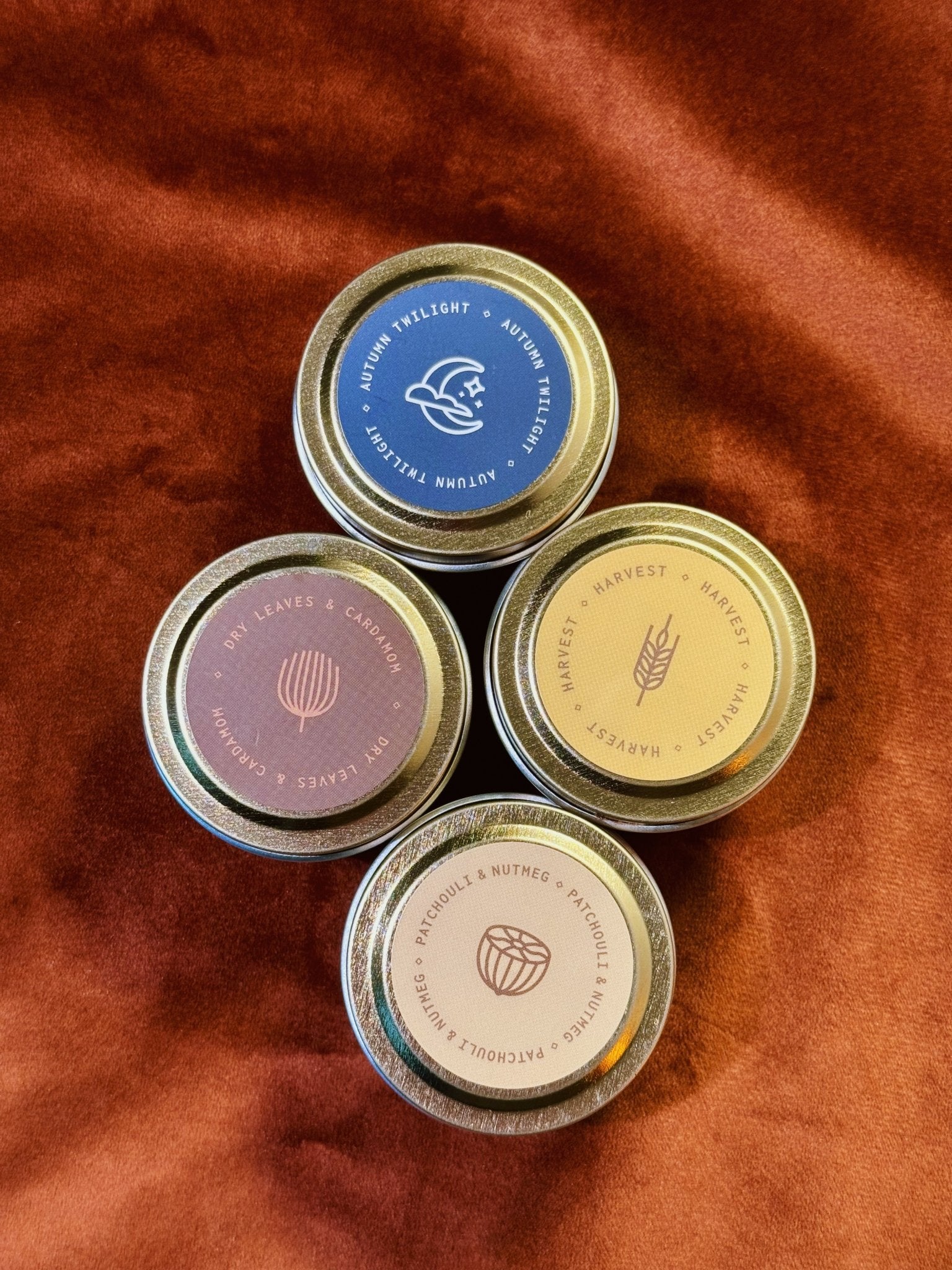 Four 2 oz tin sample candles — Autumn Twilight, Dry Leaves Cardamom, Harvest, Patchouli Nutmeg — on burnt orange fabric