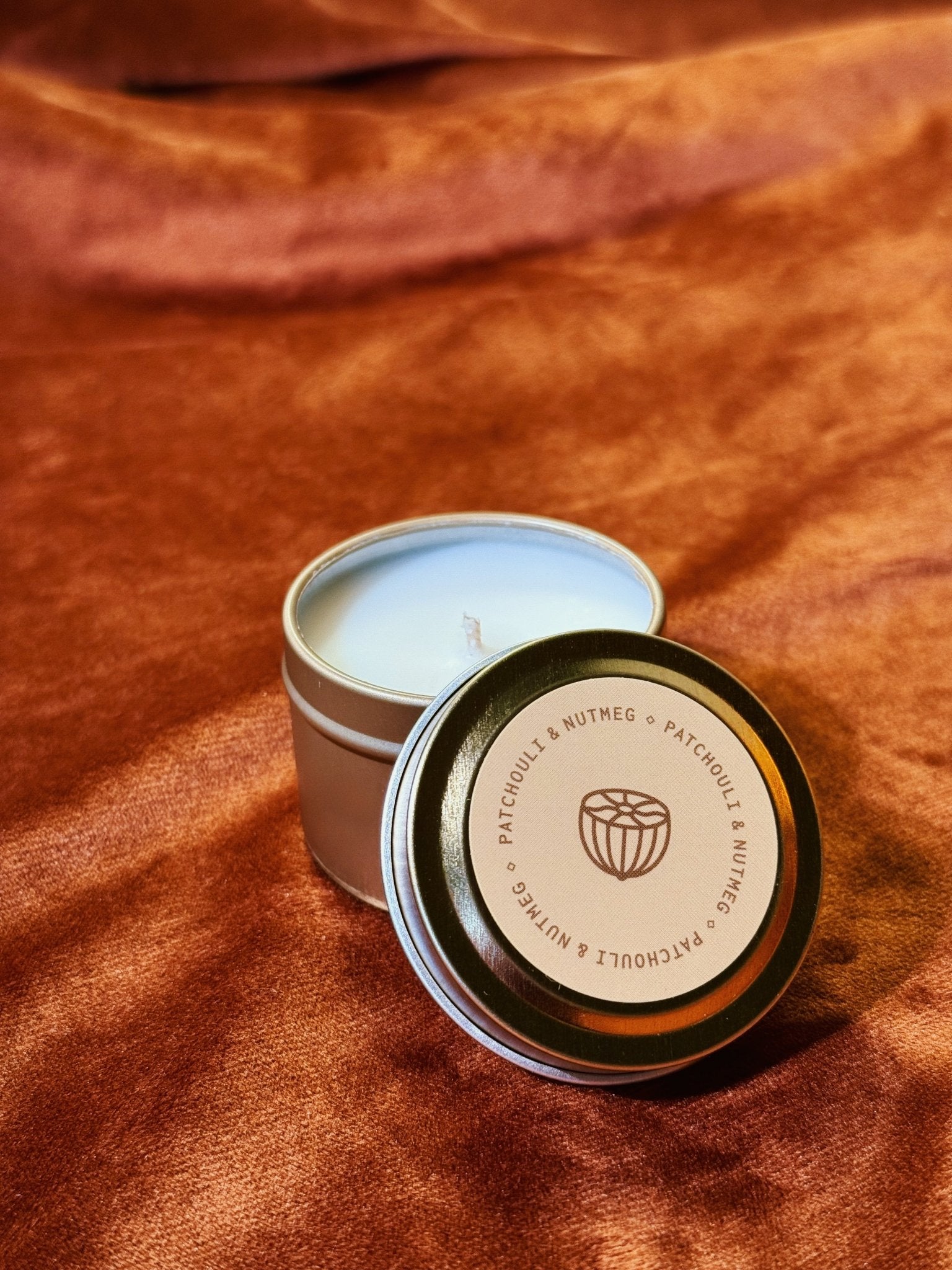 Patchouli Nutmeg 2 oz tin sample candle with taupe lid on burnt orange fabric background