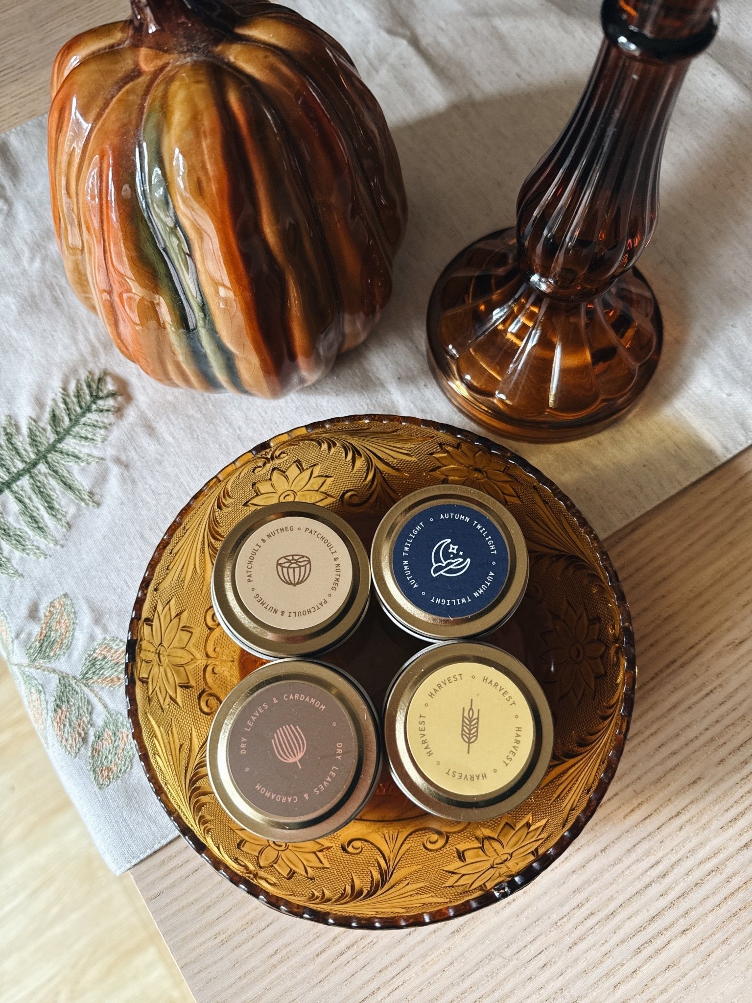 Four sample-sized autumn tin candles with lids on a decorative glass plate, surrounded by  pumpkin and a glass container