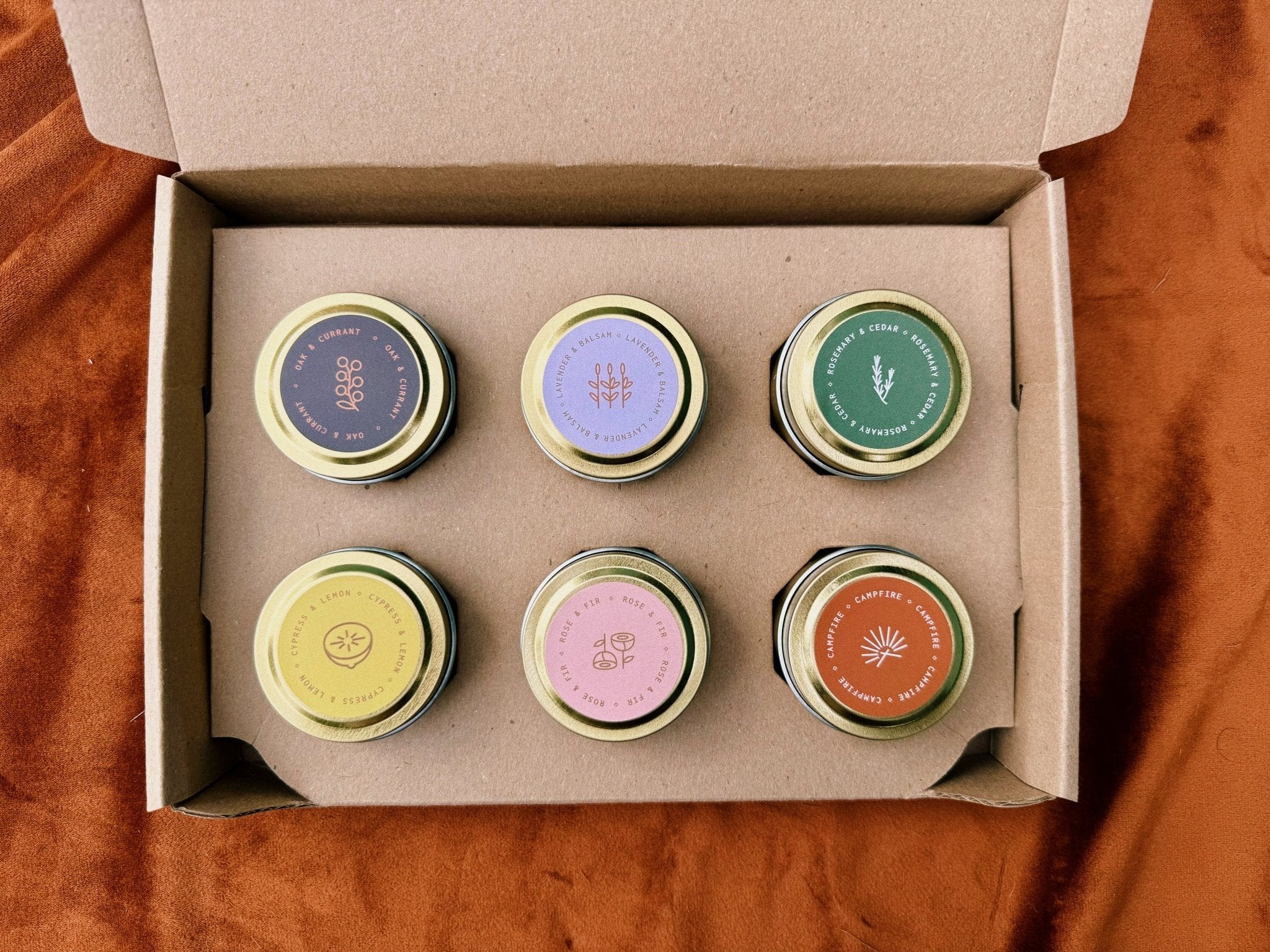 Six small candle samples with colorful lids in a cardboard box on a wooden surface