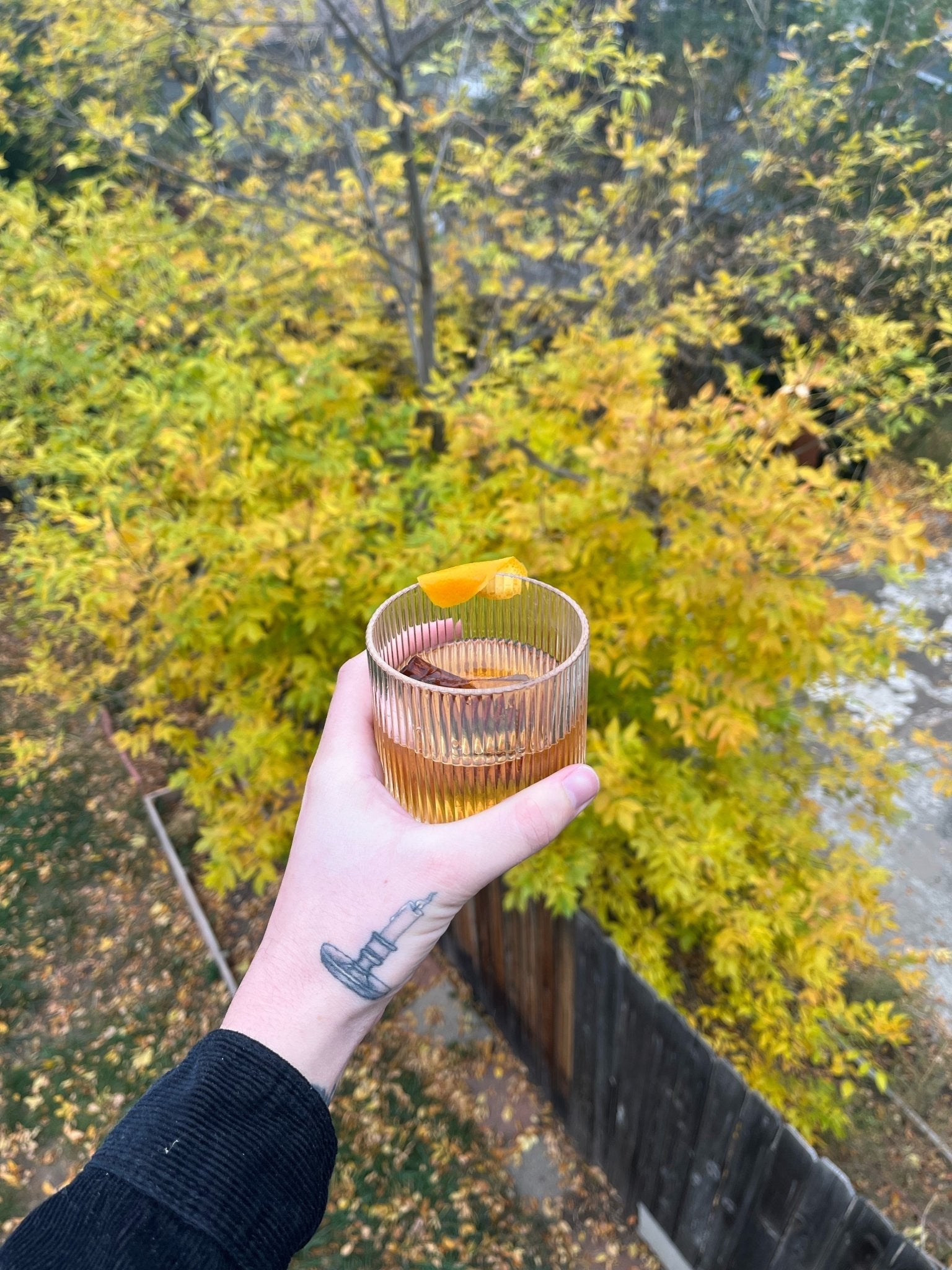 Hand holding a ribbed glass tumbler with a golden cocktail and orange peel garnish, photographed above autumn trees.