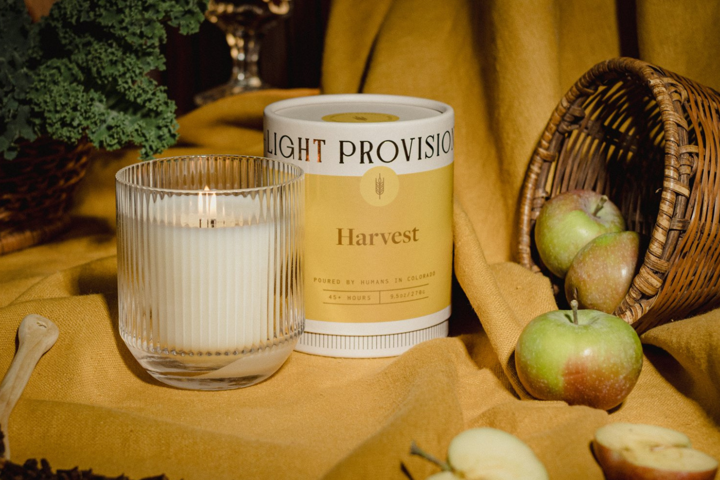 A lit candle in a ribbed clear glass holder stands beside a cylindrical tin labeled Light Provisions 'Harvest'. The scene is styled with a mustard-yellow fabric backdrop, green apples, a wicker basket, and leafy greens in a warm rustic setting.