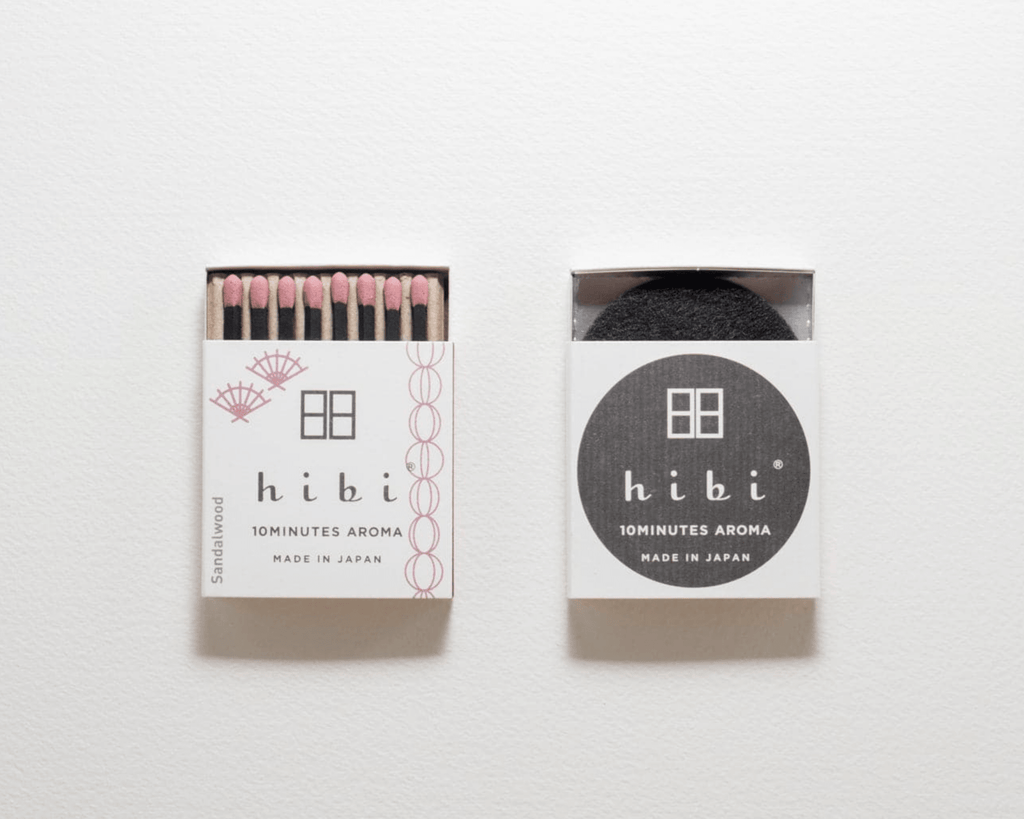 Hibi 10Minutes Aroma Sandalwood matches in white packaging on a clean white background. One matchbox is open, showing pink-tipped matches with black handles, and the other is closed with a black circular logo and a dark round insert visible inside.