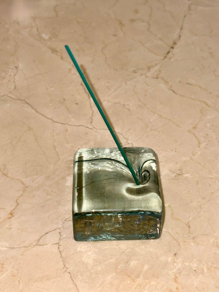 Square recycled glass reborn incense holder with a single green incense stick on a beige marble surface