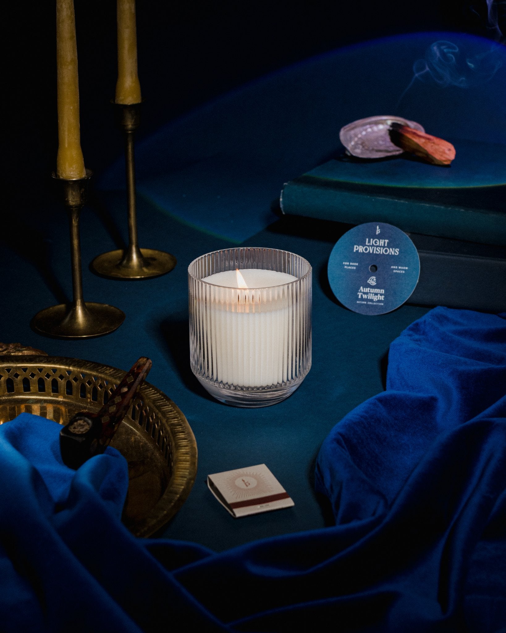 autumn twilight candle in glass tumbler sits on blue velvet surface with moody brass beeswax and ornaments. palo santo and tobacco pipe burning in background. 