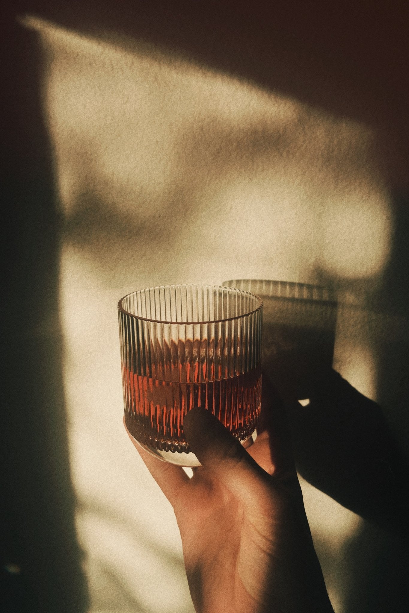 Ribbed glass tumbler with Negroni cocktail, held in one hand in warm window light with soft shadows on the wall.