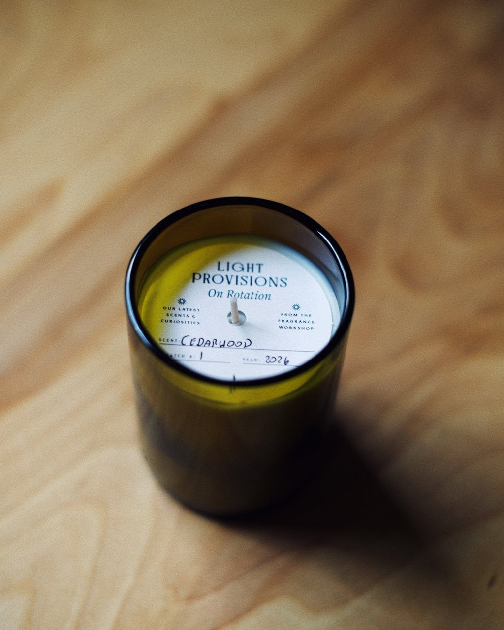 A dark amber glass jar candle with a white label reading 'LIGHT PROVISIONS On Rotation' sits on a warm-toned wooden surface.