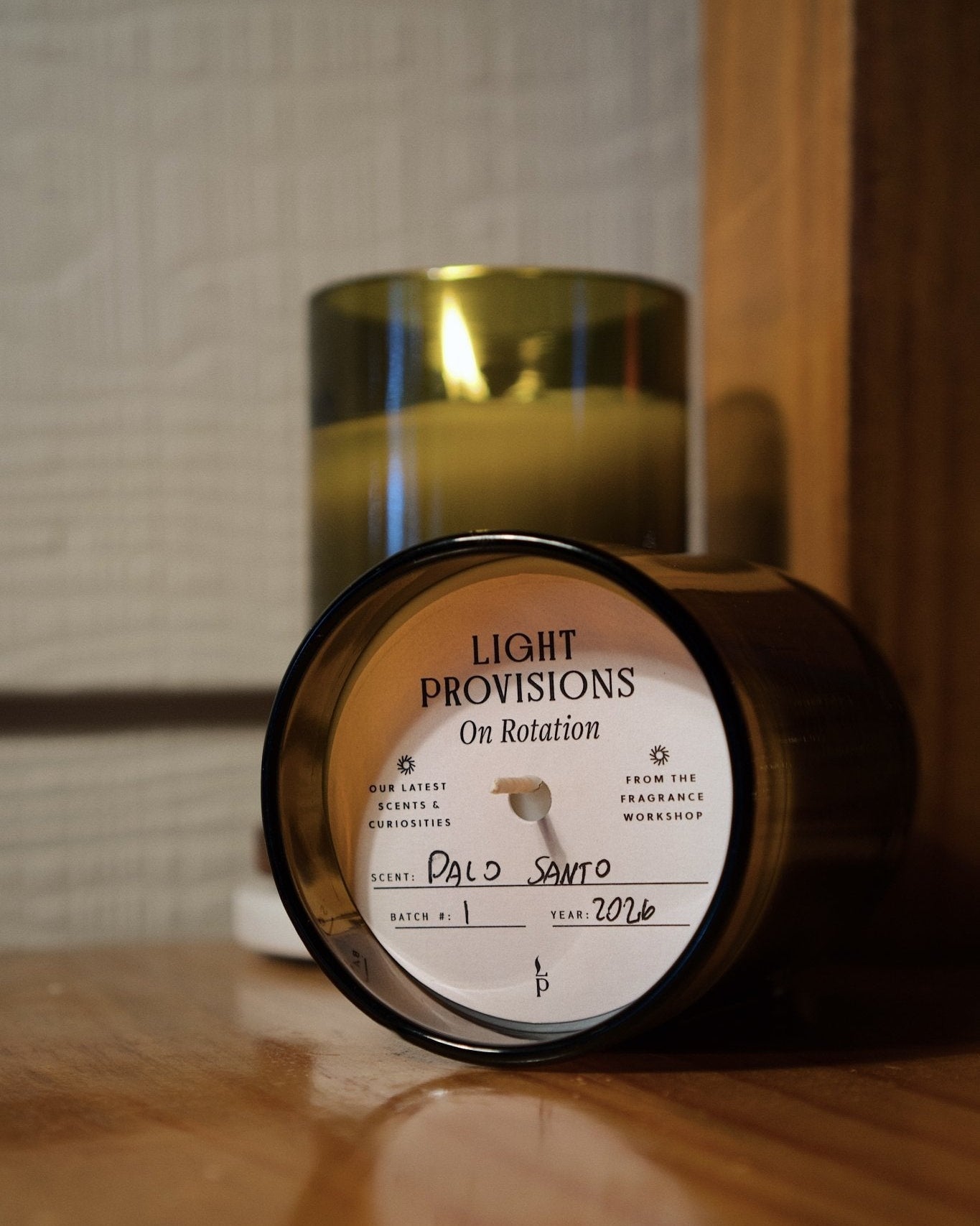 A dark amber glass jar candle lies on its side on a wooden surface, showing the front label that reads 'LIGHT PROVISIONS On Rotation' and 'Palo Santo'. A second candle stands upright in the background in a matching antique green recycled glass container.