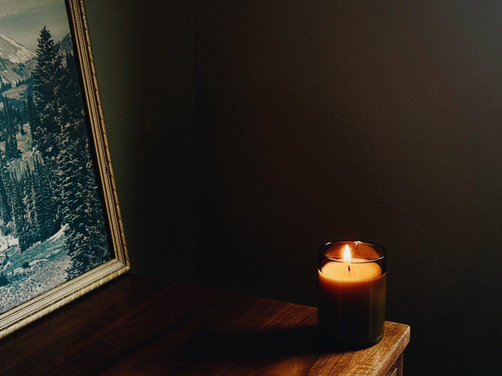 A lit candle in a amber recycled glass sits on a wooden table in a dimly lit room, next to a framed landscape print.