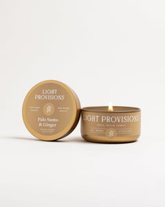 Palo Santo & Ginger small-batch travel tin candle lit, gold tin and sand label, on white background.