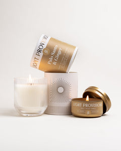 Set shot: lit ribbed-glass tumbler candle and lit gold travel tin with packaging, on white background.