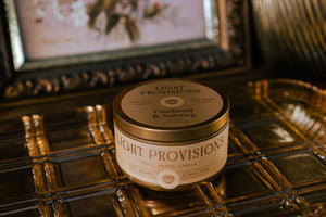 Light Provisions Patchouli & Nutmeg travel tin candle with gold lid on gold tray with framed floral art.