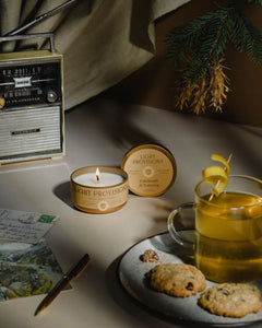 Patchouli & Nutmeg lit travel tin candle, tea, cookies and pine branch on cozy tabletop setting