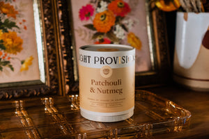 Light Provisions Patchouli & Nutmeg soy candle in glass tumbler on gold tray with floral art in background.