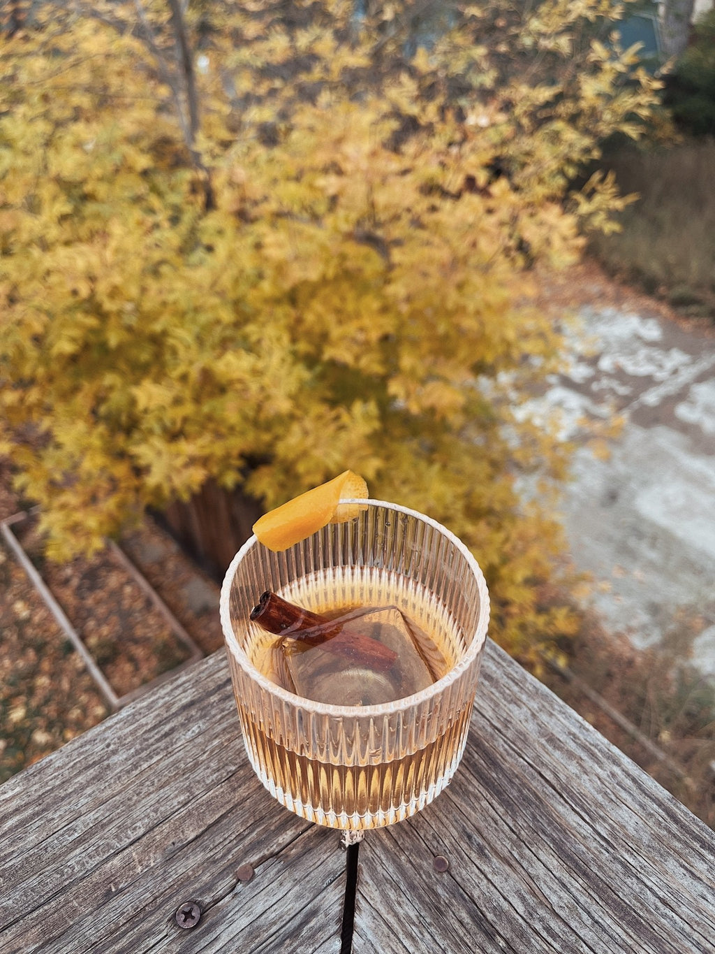 Ribbed glass tumbler filled with a bourbon based cocktail, with an orange peel, photographed outdoors above fall foliage.