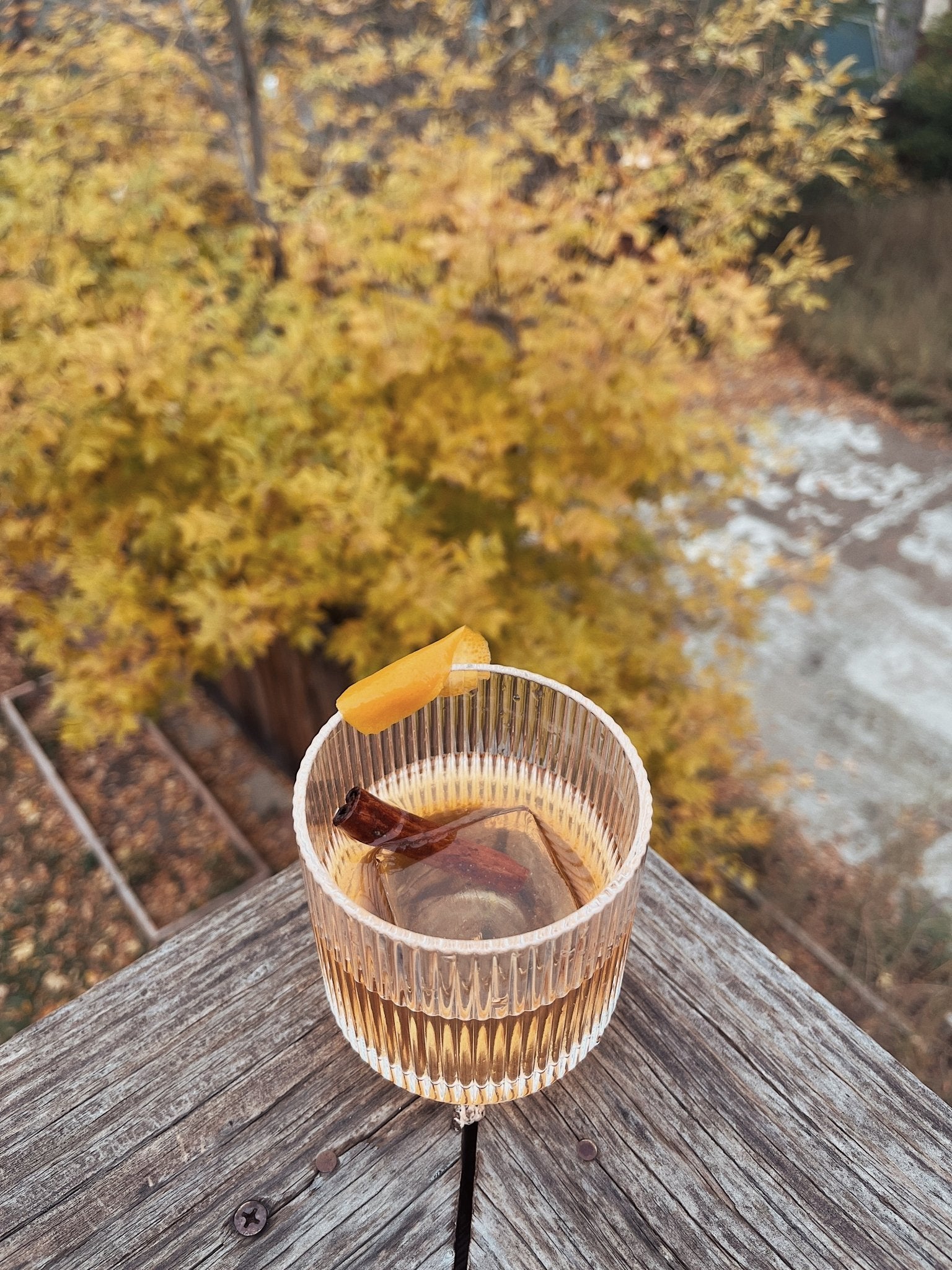 Ribbed glass tumbler filled with a bourbon based cocktail, with an orange peel, photographed outdoors above fall foliage.
