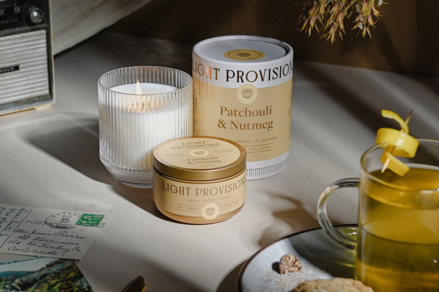 Two Light Provision scented candles in round containers, one larger tin labeled Patchouli & Nutmeg and one smaller tin, styled on a tabletop with a lit white candle in a ribbed glass holder, a glass of yellow tea, and warm ambient lighting.