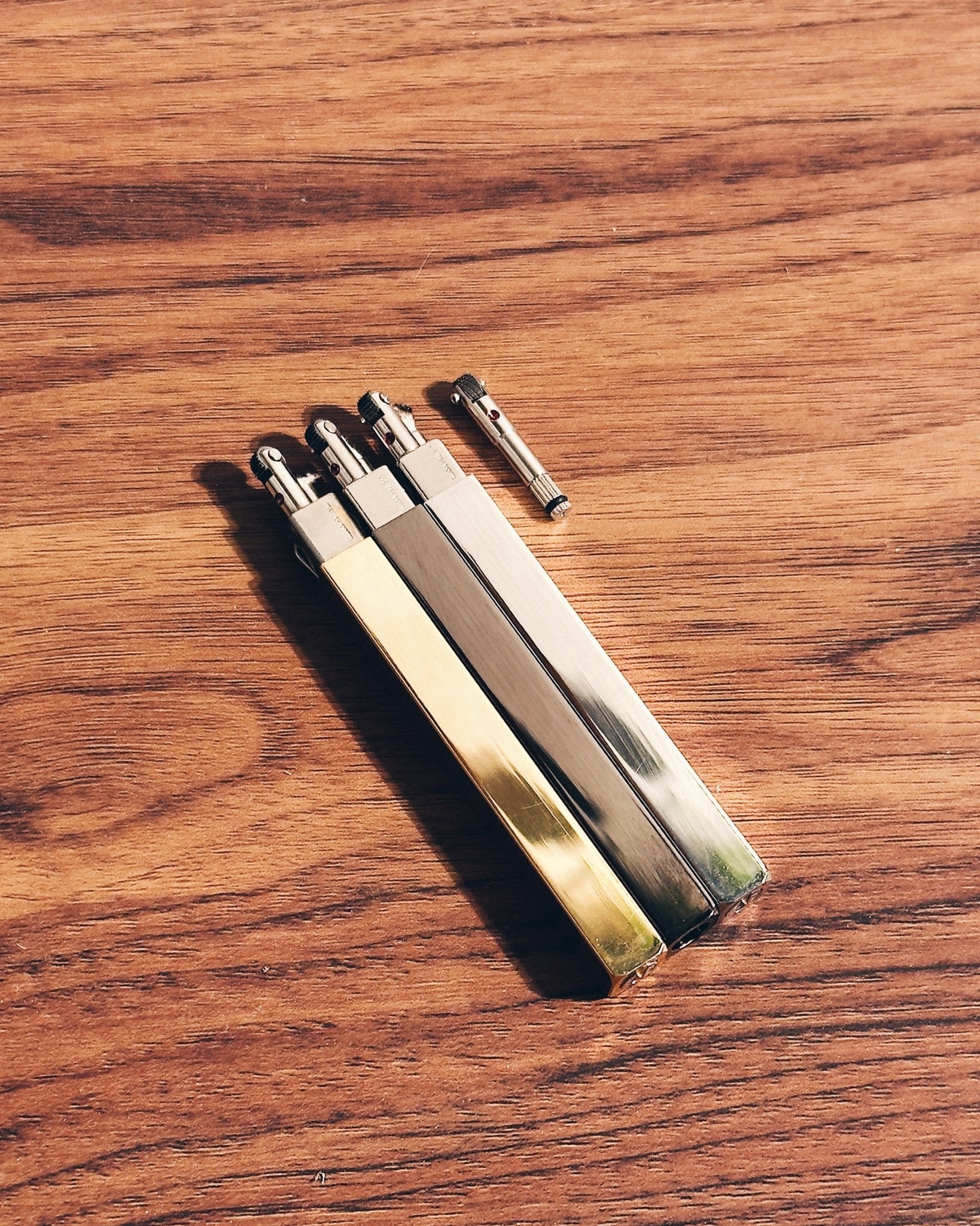 Three Tsubota Pearl Queue lighters in gold, silver, and black next to flint assembly replacement part