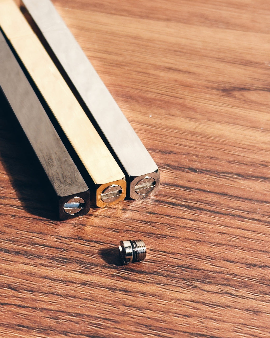 Three Tsubota Pearl lighters in black, gold, and silver finishes with a small metal threaded screw, arranged on a wooden surface.