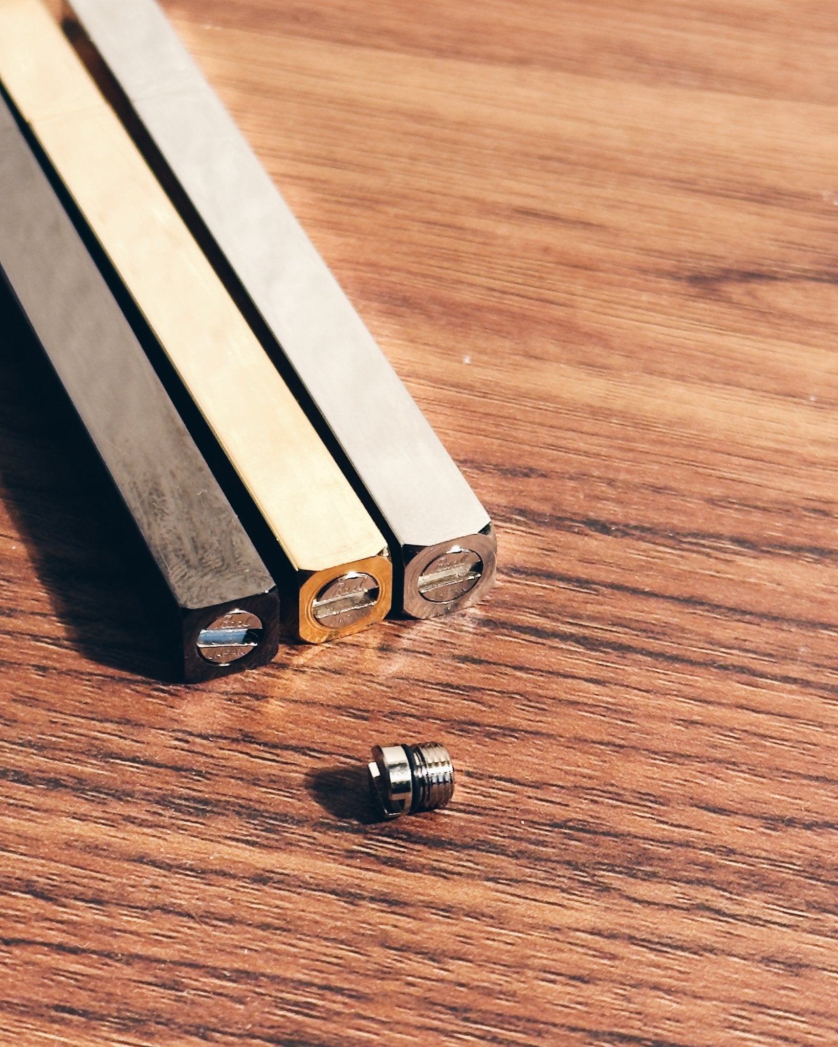 Three Tsubota Pearl lighters in black, gold, and silver finishes with a small metal threaded screw, arranged on a wooden surface.