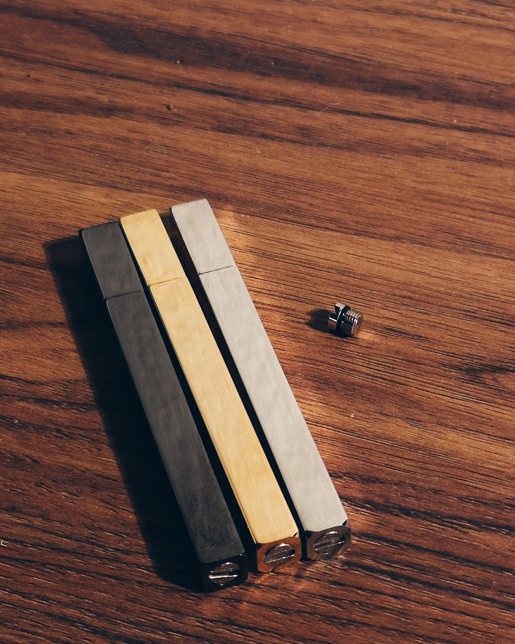 Three Tsubota Pearl lighters in black, gold, and silver finishes with a small metal threaded screw, arranged on a wooden surface.