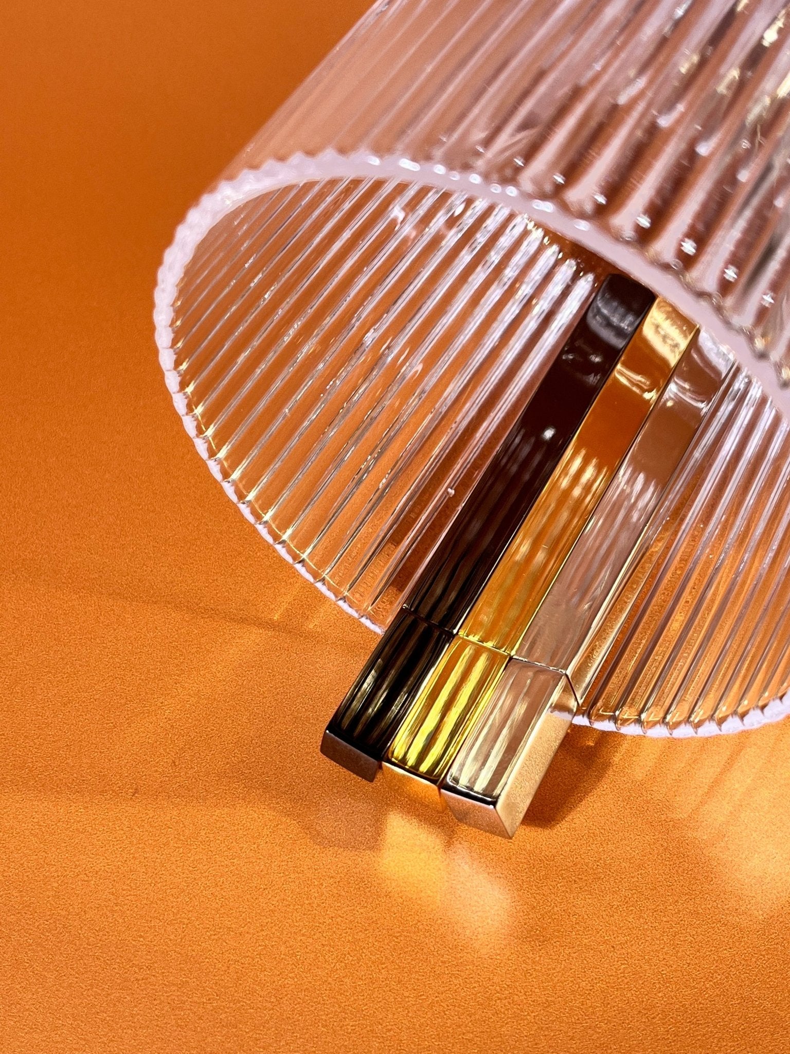 Three slim Queue stick lighters in silver, gold, and brass finishes styled under a ribbed glass tumbler on orange background