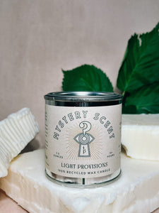 Candle labeled 'Mystery Scent' from 'Light Provisions' on a beige background with green leaves and soy wax chunks