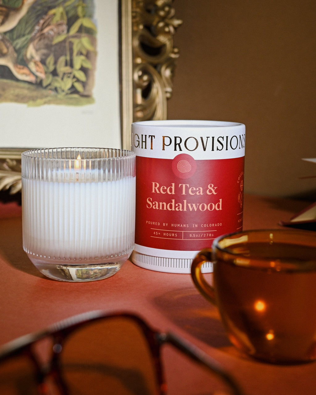 Red Tea & Sandalwood glass tumbler candle beside matching tin and amber cup, reflection visible on burnt orange surface.