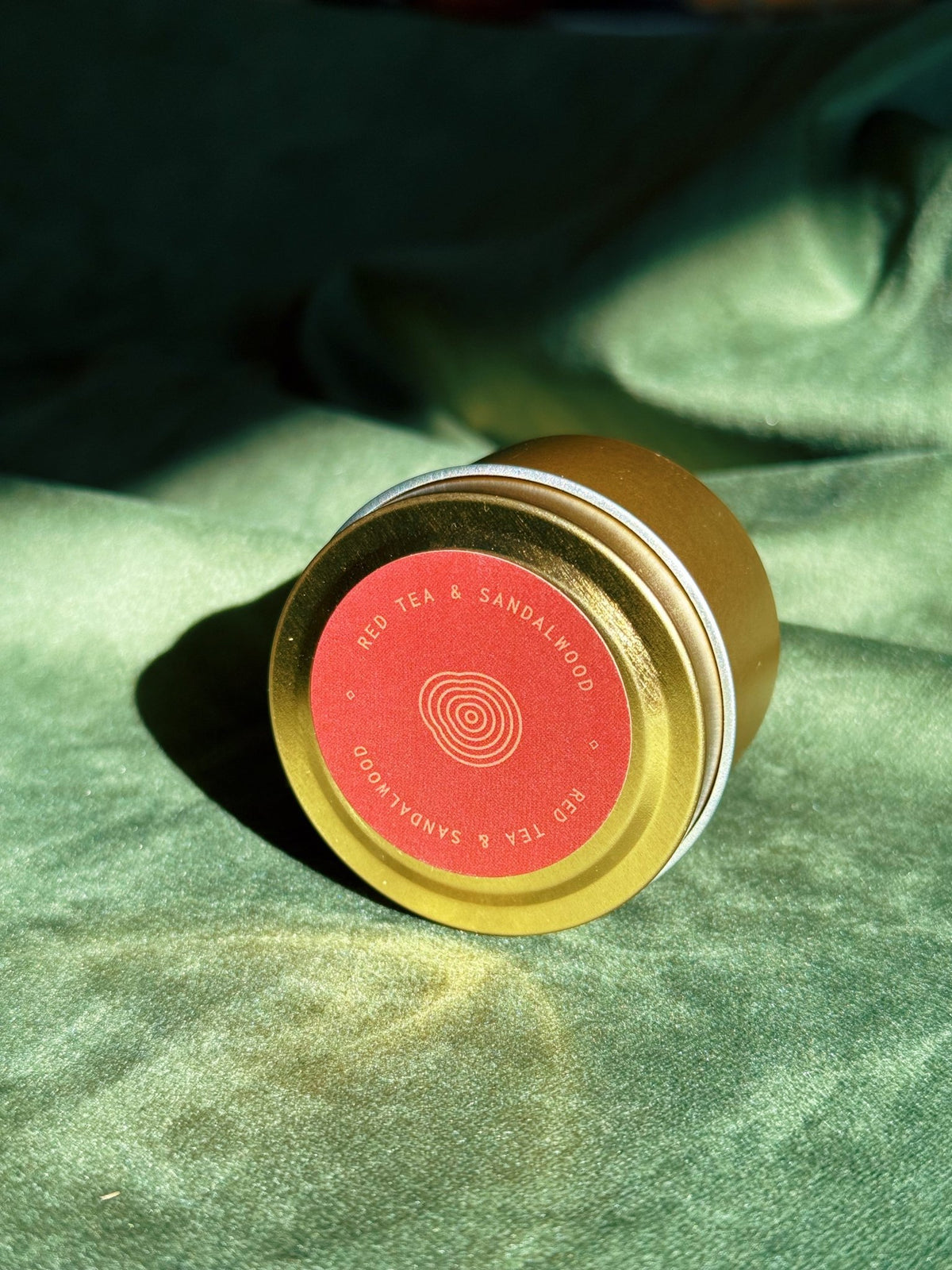 Gold sample tin labeled Red Tea and Sandalwood resting on green fabric with warm sunlight.
