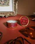 Red Tea & Sandalwood tin and glass candle with gold spoon and amber drinkware, warm tabletop under afternoon light.