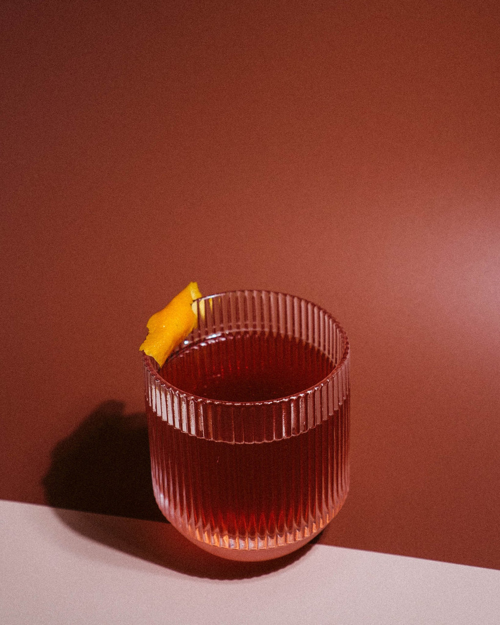 Fluted low ball glass with an old fashioned cocktail and orange peel garnish, photographed against a warm brown backdrop.