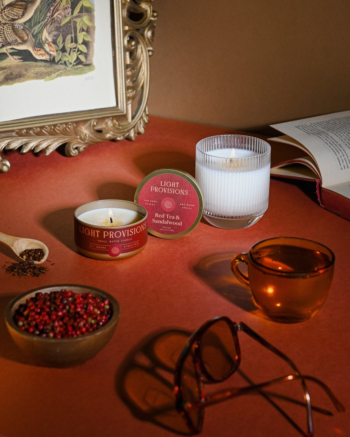 Red Tea & Sandalwood candles with peppercorns, amber glass, and vintage frame casting soft shadow across warm scene.