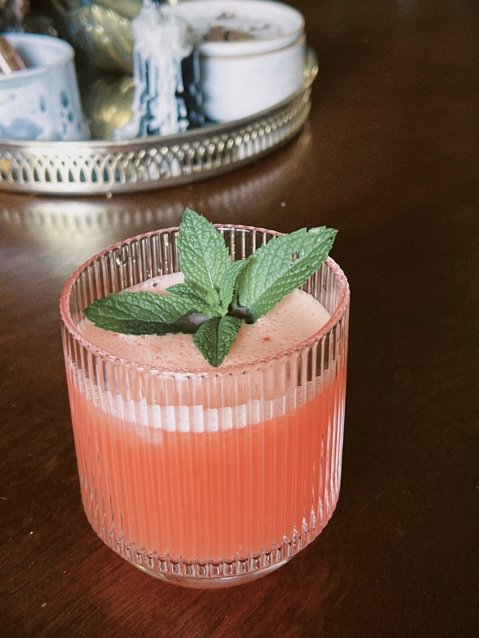 Ribbed glass with a pink cocktail topped with fresh mint leaves, photographed on a dark wooden table.