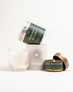 Set shot with lit ribbed-glass tumbler candle and lit Rosemary & Cedar travel tin, packaging behind, on white background.