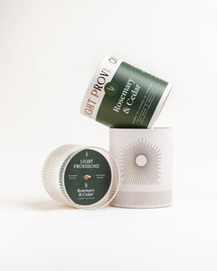 Styled display: tilted Rosemary & Cedar tumbler box and sunburst canister next to ribbed glass lid, on white background.