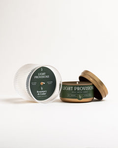 Open Rosemary & Cedar gold travel tin beside ribbed glass holder lid, unlit wick, on white background.