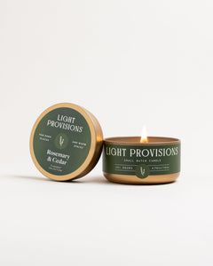 Rosemary & Cedar small-batch travel tin candle lit, gold tin with green label, on white background.