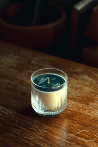Single fluted tumbler resting on wood; dark background; top reads “LIGHT PROVISIONS · Rosemary & Cedar,” wick centered
